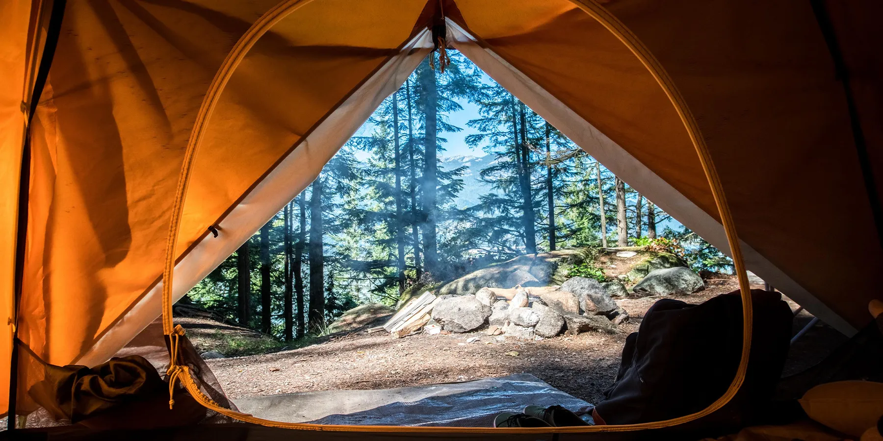 View from inside an orange tent looking out at a forest campsite with a smoking fire ring and tall pine trees