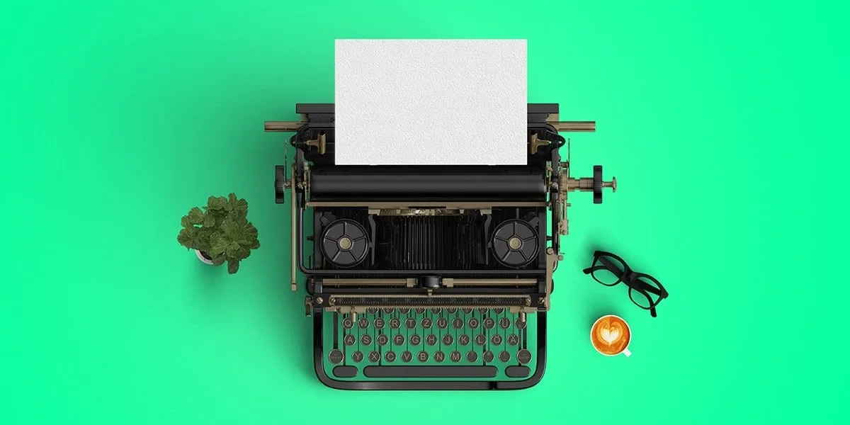 Overhead flat-lay of a vintage typewriter with blank paper, a potted plant, glasses and a heart-shaped latte on bright green