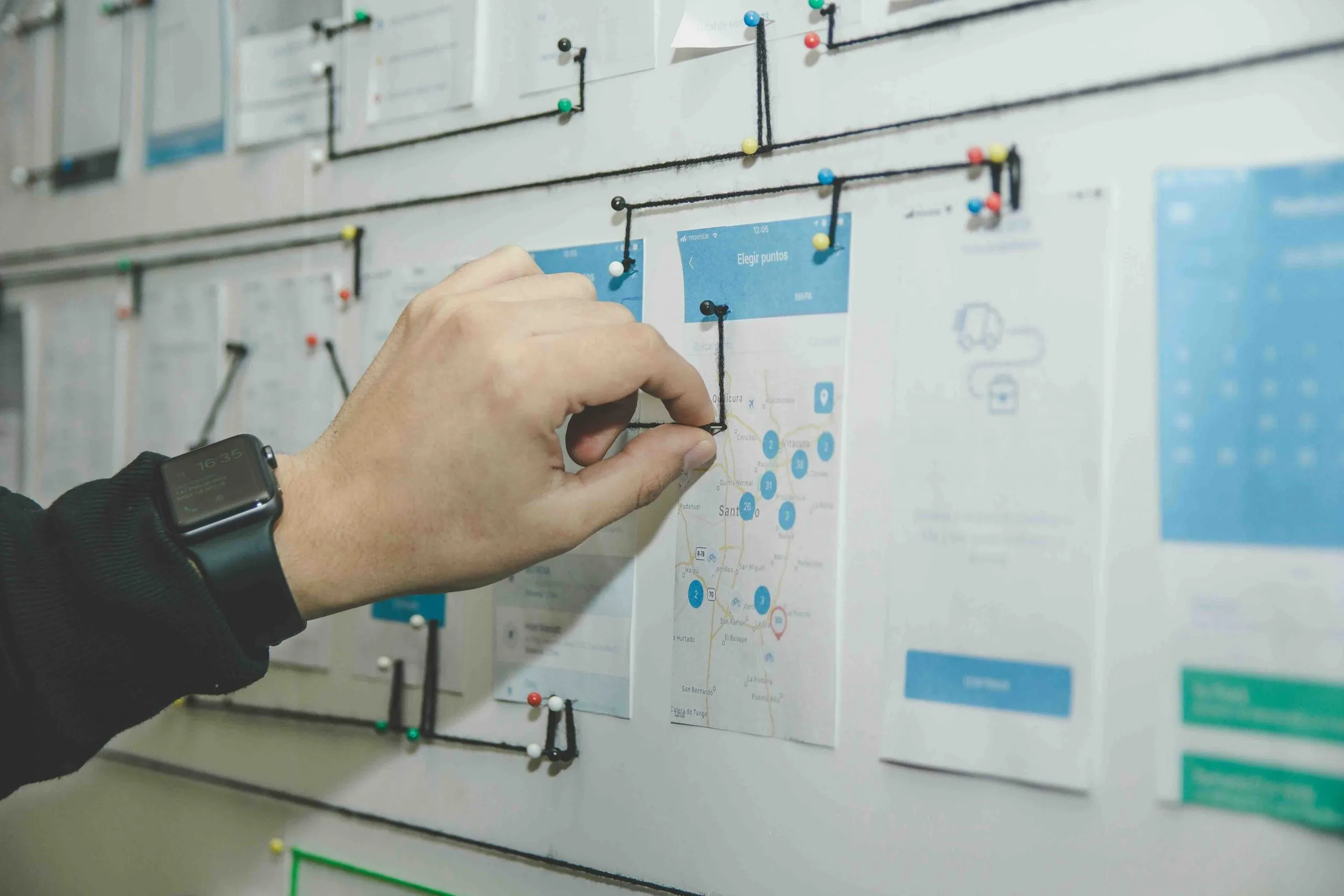 A hand pins black string between printed mobile app wireframes on a whiteboard, mapping out a user-flow diagram