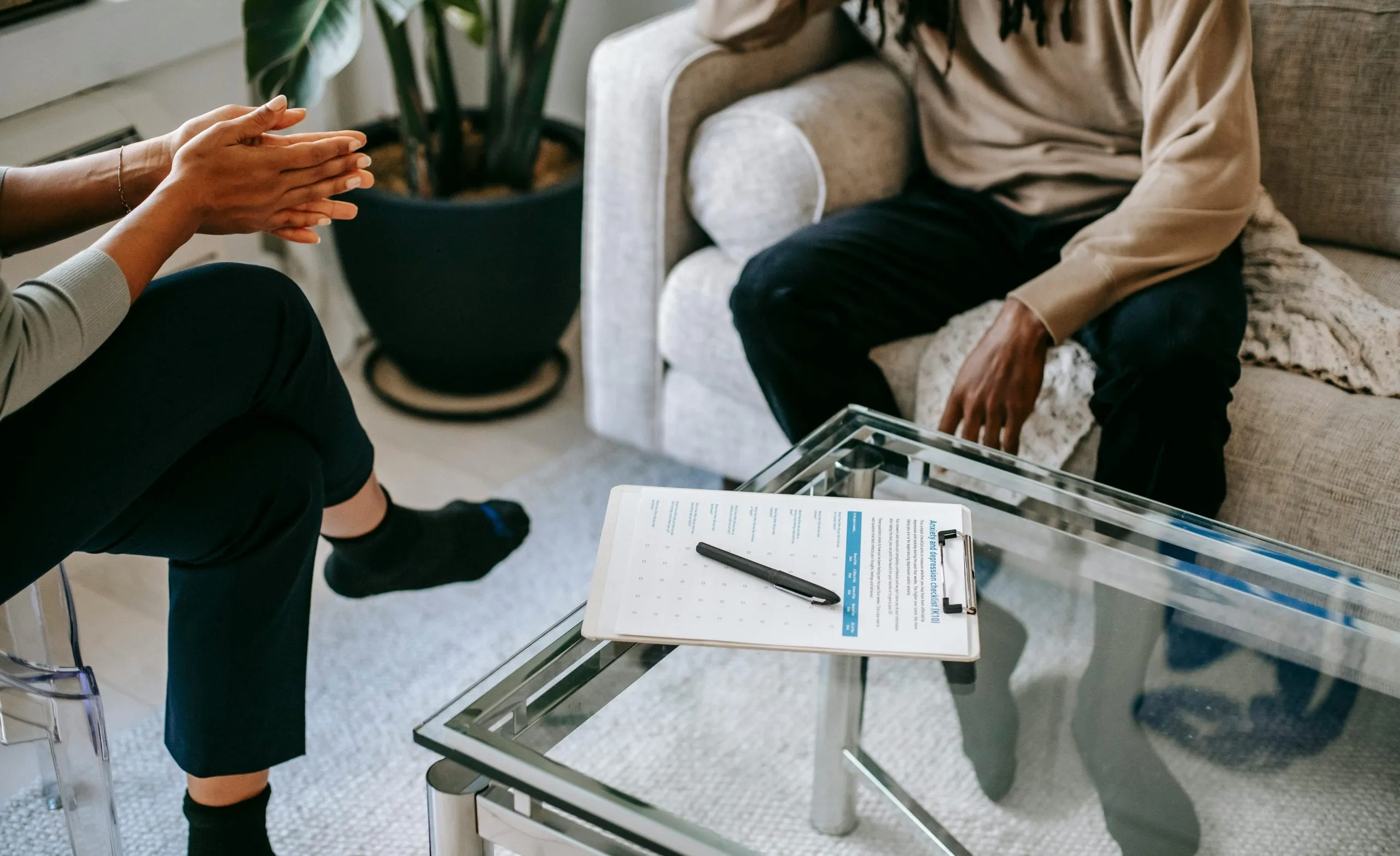 Therapy session with a client seated on a couch and a counselor across a glass table holding an assessment clipboard