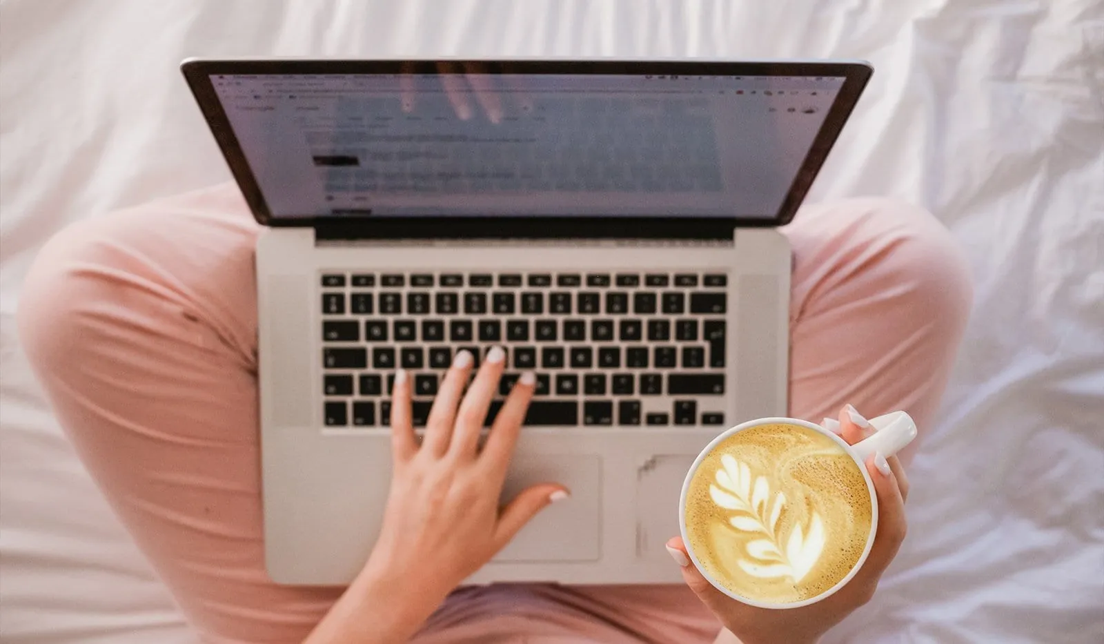 Overhead view of a person in pink pajamas typing on a laptop in bed while holding a cappuccino with leaf latte art