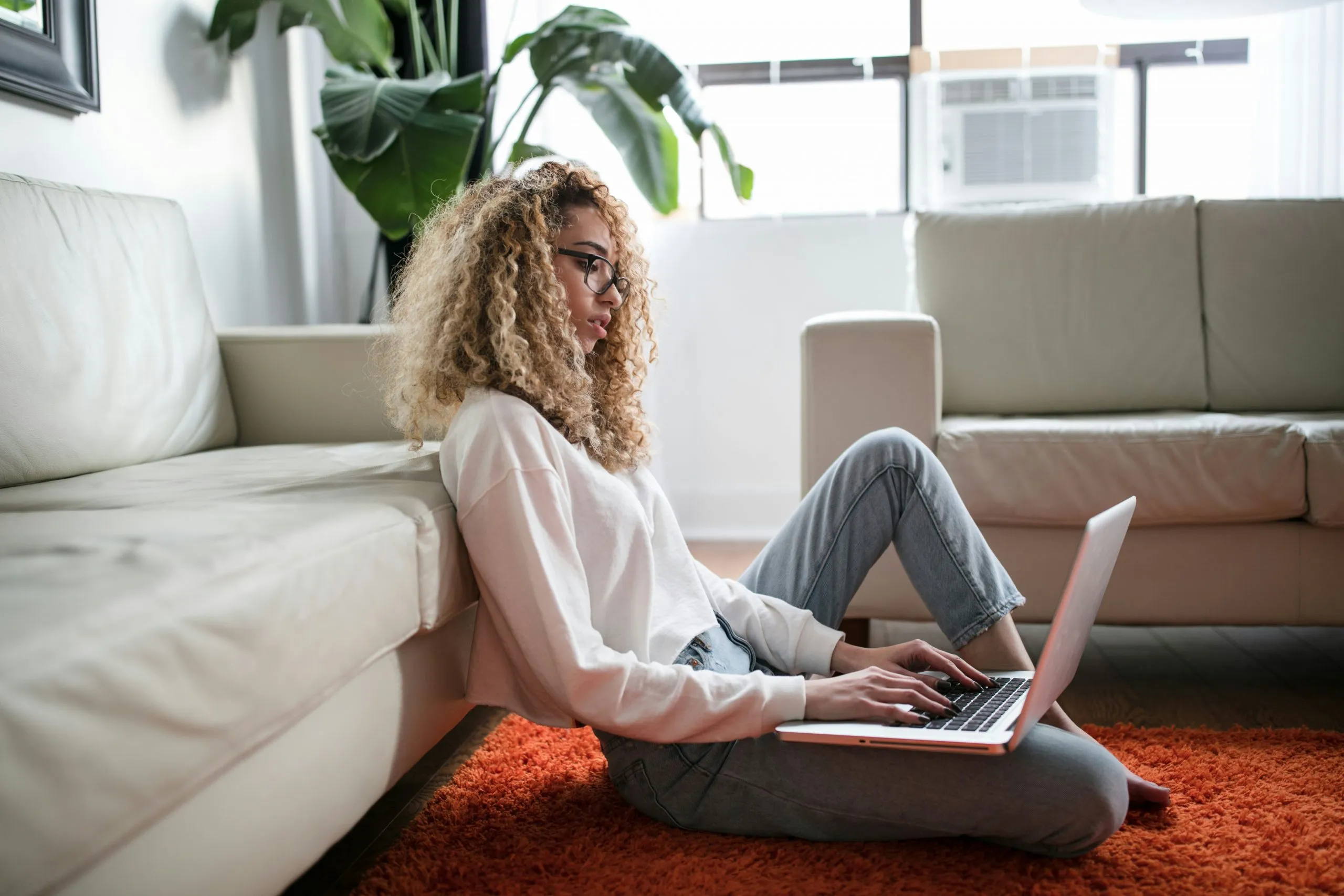Young woman with curly hair and glasses sitting on the floor against a couch, typing on a laptop in a sunlit room