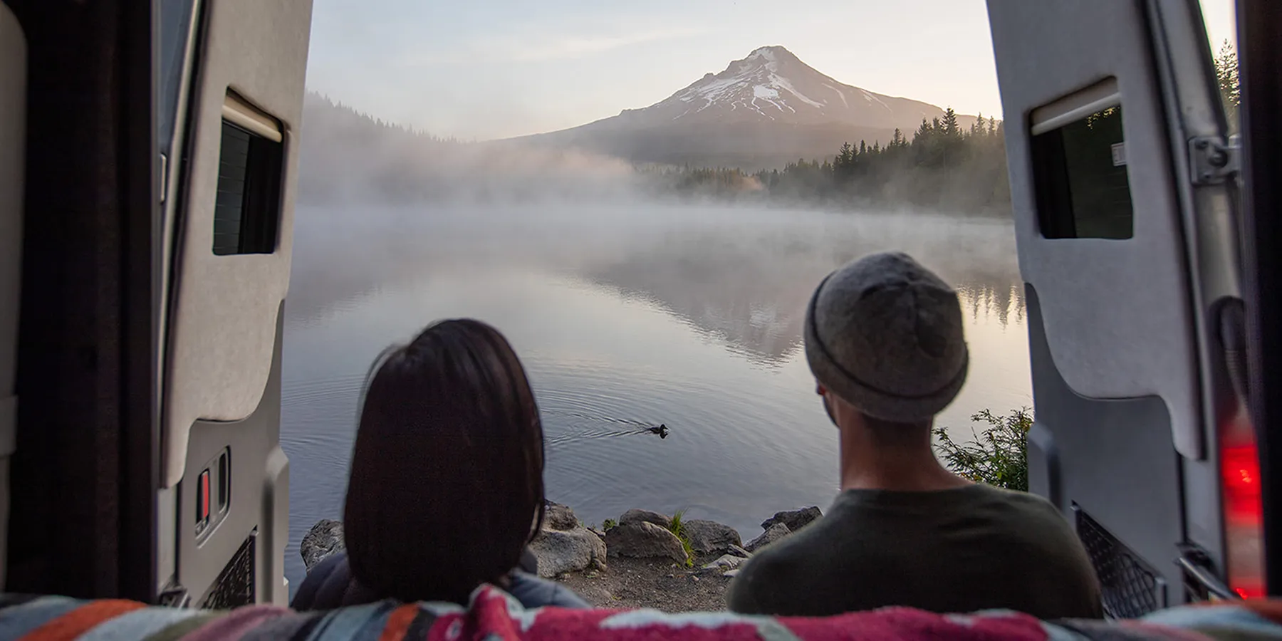 Couple seen from inside a camper van looking out the open rear doors at a misty lake and snow-capped Mount Hood at dawn