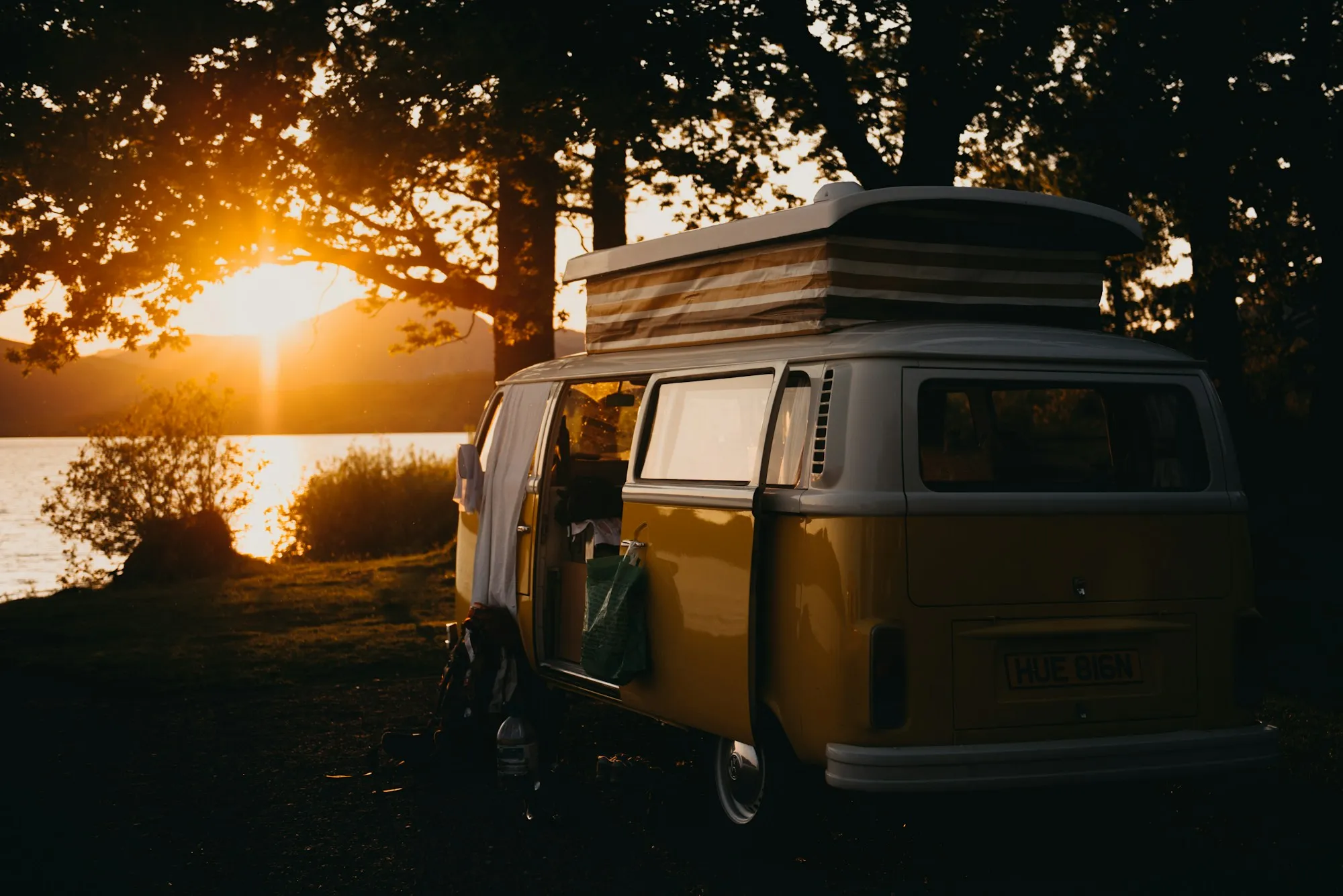 Yellow vintage VW camper van with pop-top roof parked by a lake at sunset, doors open and a backpack on the ground