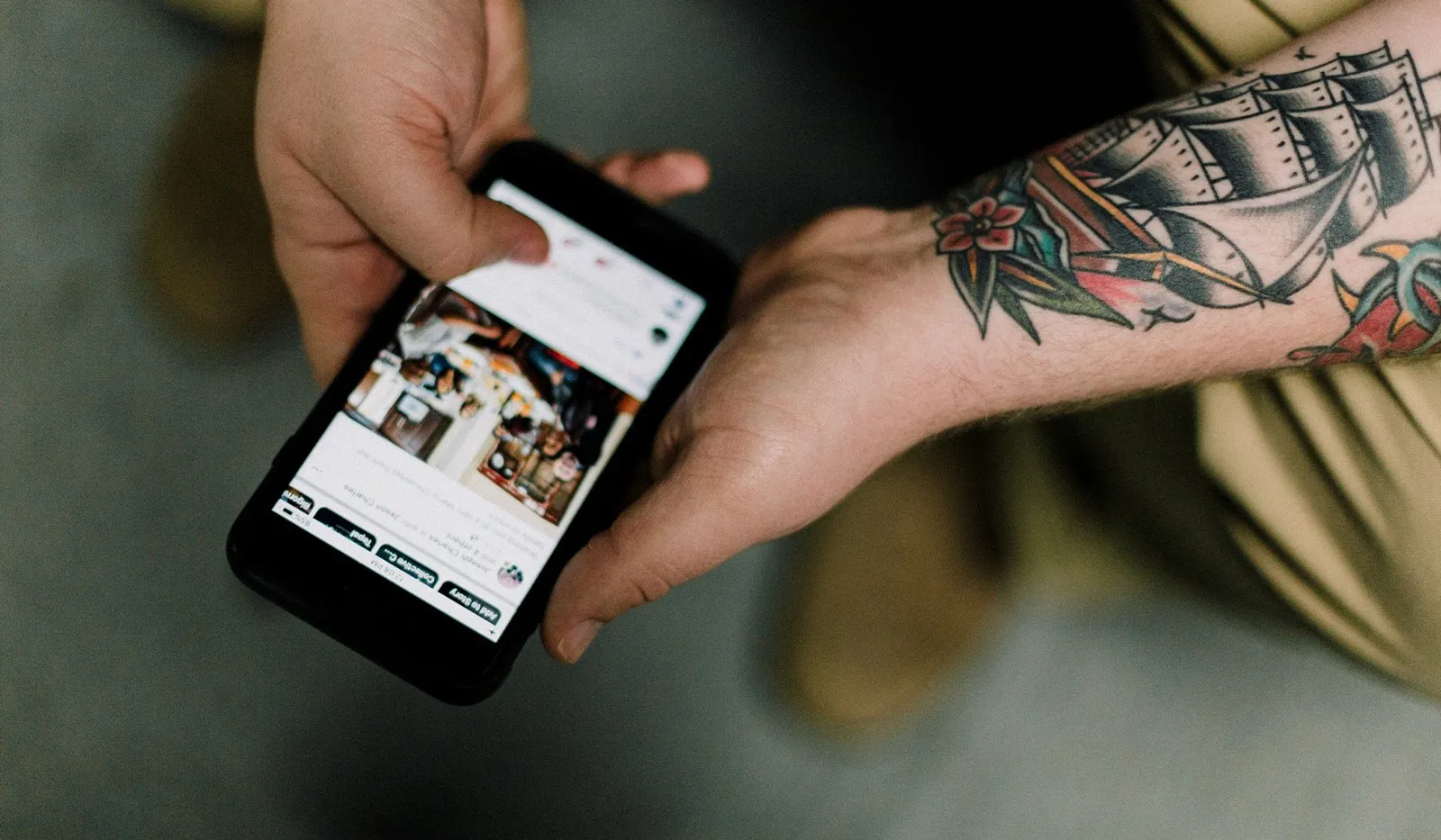 A tattooed arm holds a black smartphone showing a social media feed, with a ship tattoo visible on the forearm