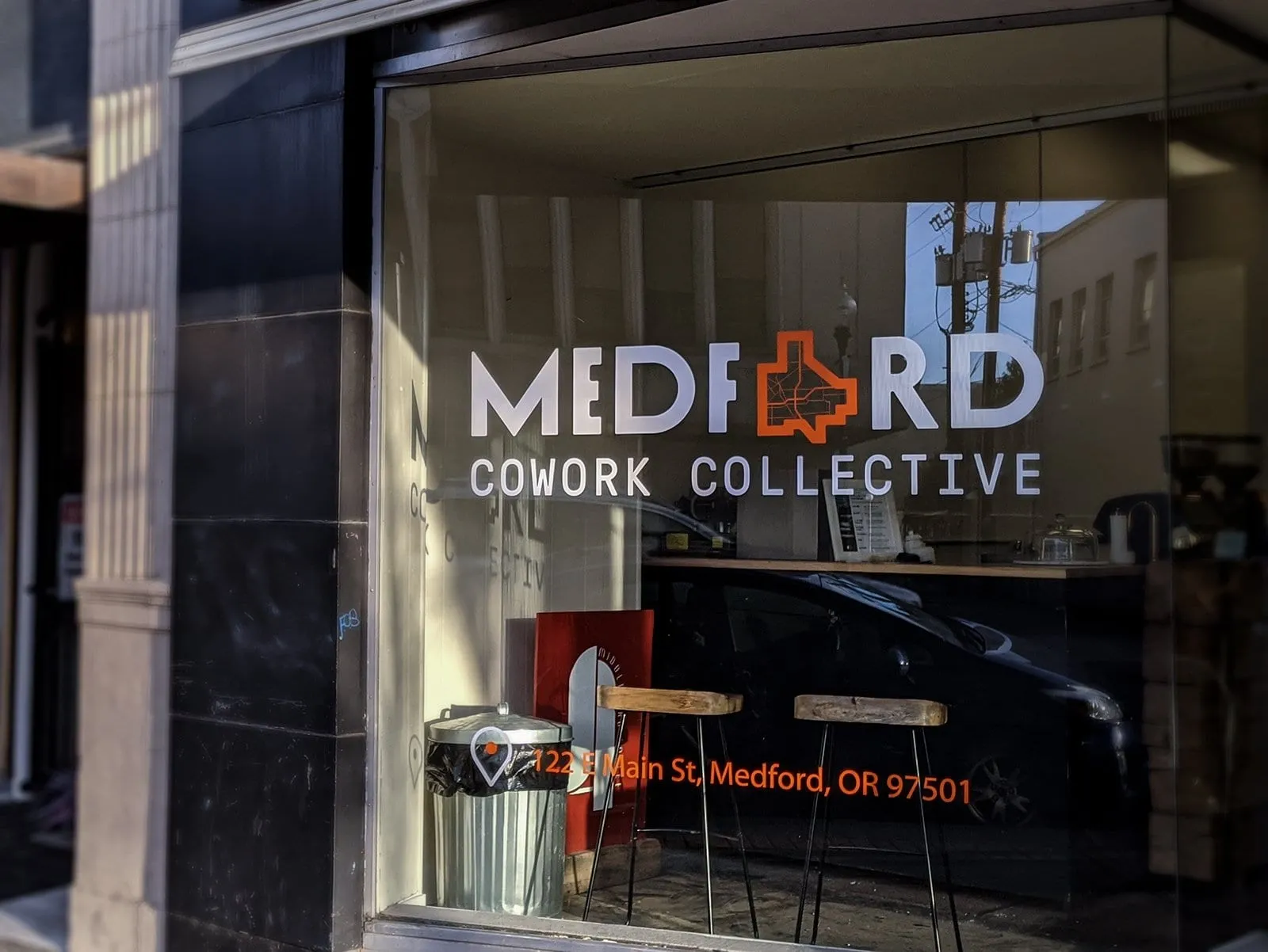Storefront window with 'Medford Cowork Collective' lettering and logo, bar stools and the street reflected in the glass
