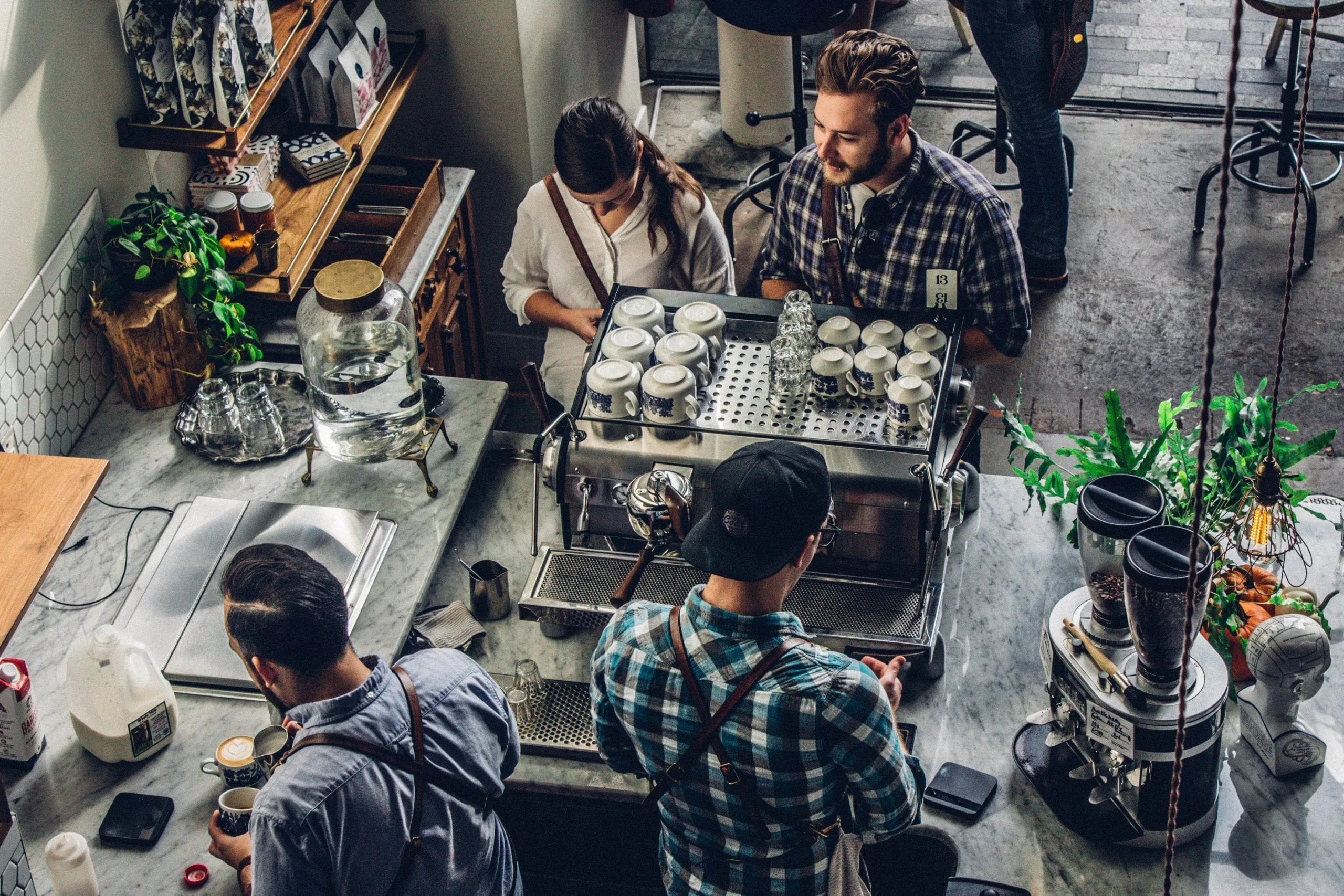 Overhead view of customers ordering at a cafe counter while baristas work behind an espresso machine stacked with cups