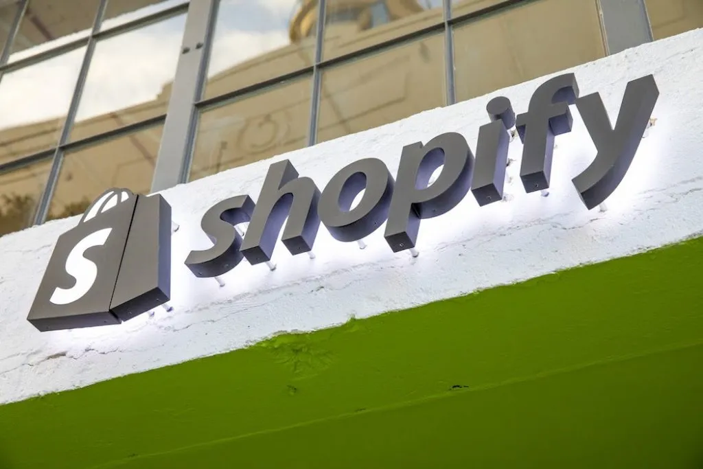 Illuminated Shopify storefront sign with shopping-bag logo mounted on a white-and-green building exterior