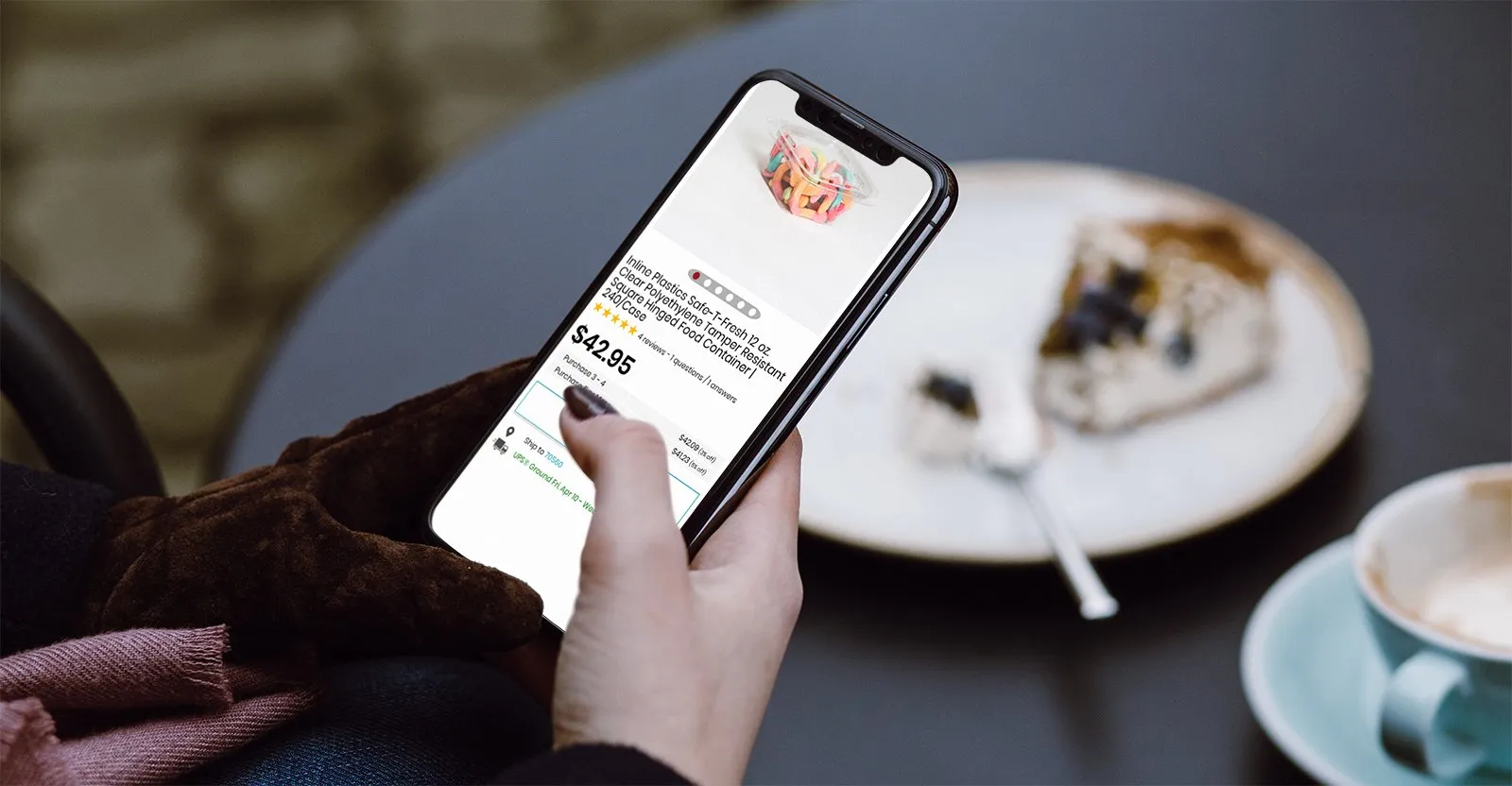 Gloved hand holding a phone showing a PJP food-container product page priced $42.95, beside coffee and dessert at a cafe