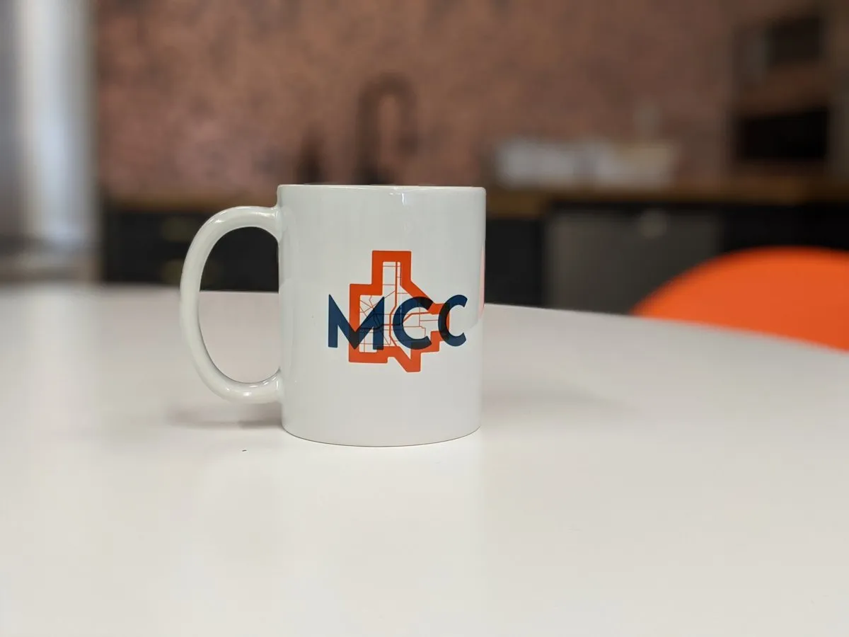 Branded white coffee mug showing the 'MCC' monogram over the orange-and-blue city-map mark